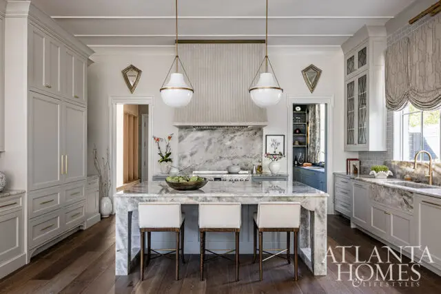 Bright kitchen with white cabinetry, marble island, brass fixtures, and woven window shades.