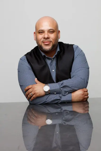 Man with dark hair and beard, wearing a vest and shirt, arms crossed.