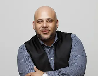 Man with short dark hair and beard, wearing a blue shirt and black vest, arms crossed.