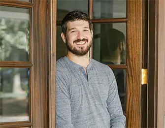 Man with beard smiles, leaning against a wooden door with glass panes.