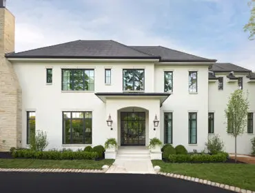 Two-story white brick home with stone chimney and black-framed windows, landscaped front yard.
