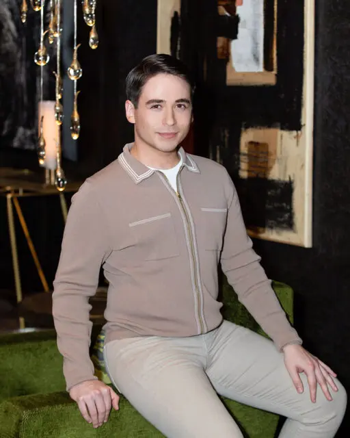 Man with short dark hair wearing a taupe zip-up jacket and light-colored pants.