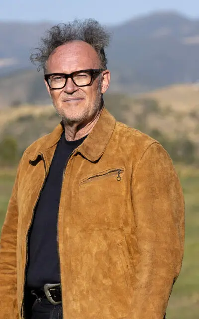 Man in a brown suede jacket stands in a grassy field with mountains in the background.