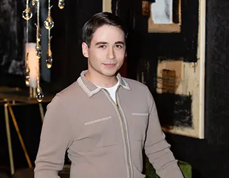 Man in a beige jacket poses with artwork and hanging crystals.