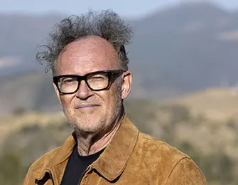 Bill Peace, wearing glasses and a suede jacket, outdoors with mountains in the background.