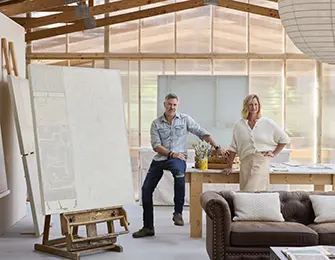Two people stand in a studio with a large canvas and furniture.