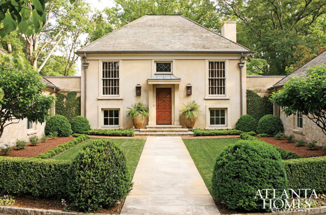 Light-colored stucco house with a gray shingle roof, tall windows, and a wooden front door. The entrance is flanked by large potted ferns and manicured hedges.