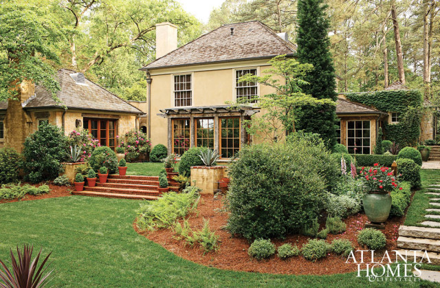 Yellow stucco house with stone accents, a shingled roof, and large windows, surrounded by lush green landscaping and mature trees.