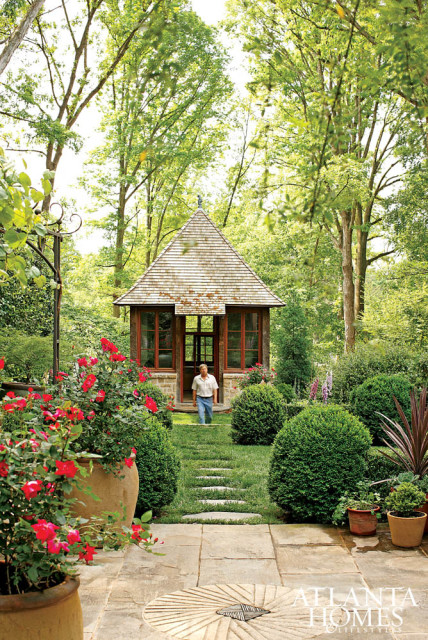 Stone and wood garden folly with a conical roof surrounded by lush greenery, manicured bushes, and blooming red roses. A man walks down a stepping stone path towards the folly.