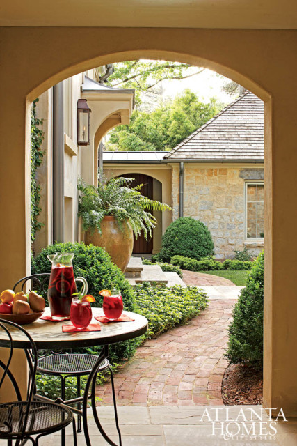 Arched entryway to a stone house with a brick pathway, lush greenery, and a table set with drinks and fruit.