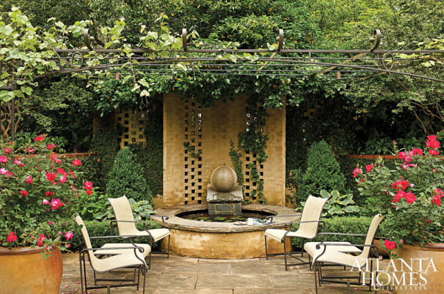 Garden patio with a circular stone fountain, a metal arbor covered in vines, and four outdoor chairs surrounded by lush greenery and pink roses.