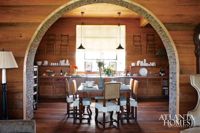 Rustic dining room with wood-paneled walls, a large wooden table set for a meal, and a stone archway leading to a kitchen area with a sink and cabinetry.