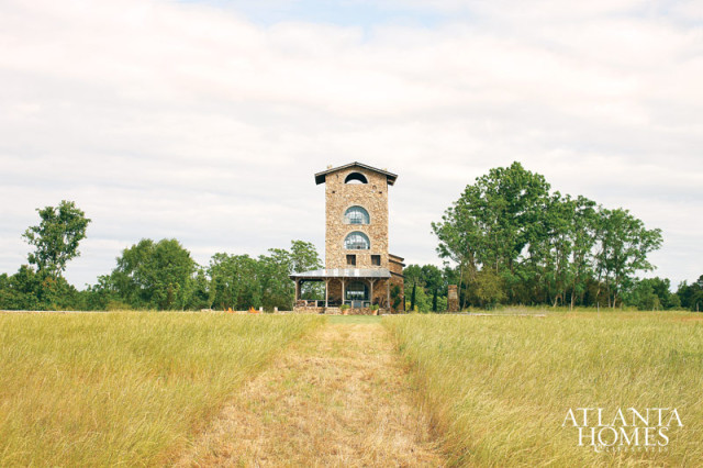 Stone tower with arched windows and a covered porch, set in a grassy field with trees under a cloudy sky. Atlanta Homes & Lifestyles logo in the lower right corner.