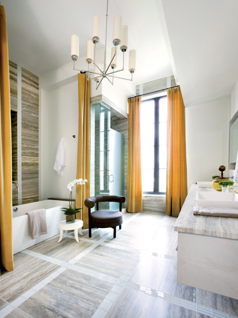 Modern bathroom with a freestanding tub, glass-enclosed shower, double vanity with marble countertop, and a dark upholstered chair. Ochre curtains frame a tall window.