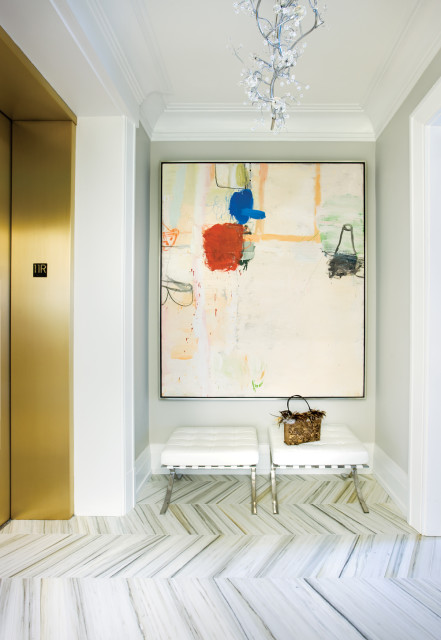Hallway with gold elevator doors, abstract art, two white tufted benches, and marble herringbone flooring.