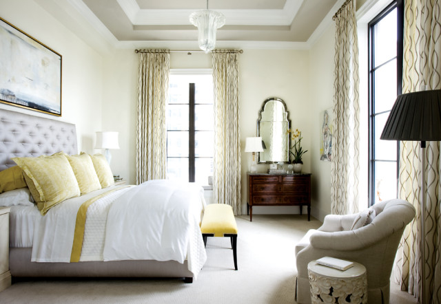 Bedroom with a tufted gray headboard, yellow accent pillows, white bedding, and patterned curtains. A dark wood dresser with a mirror sits opposite the bed.