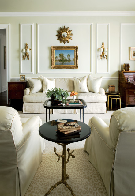 Pale green living room with a cream sofa, two armchairs, and a black coffee table with books and plants. A landscape painting hangs above the sofa.