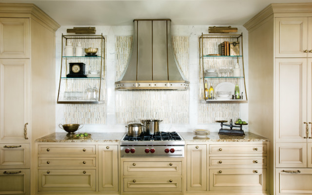 Cream kitchen with a stainless steel range hood, marble backsplash, and glass-fronted shelves displaying decorative items and dishware.
