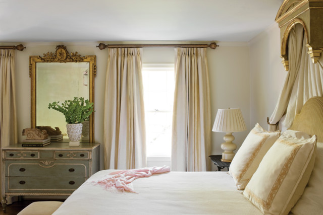 Bedroom with a light green dresser topped with a gold ornate mirror and a green plant, cream curtains, and a bed with cream pillows and a pink throw