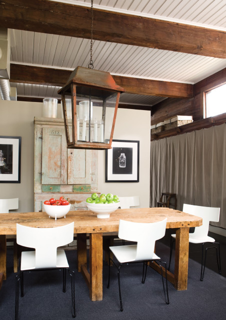 Rustic wooden dining table with white chairs, antique cabinet, and a large metal lantern chandelier. Bowls of tomatoes and green apples are on the table.