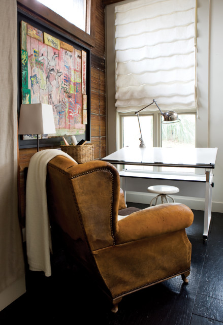 Brown leather armchair next to a desk with a white Roman shade window, artwork on wood-paneled wall, and a floor lamp.