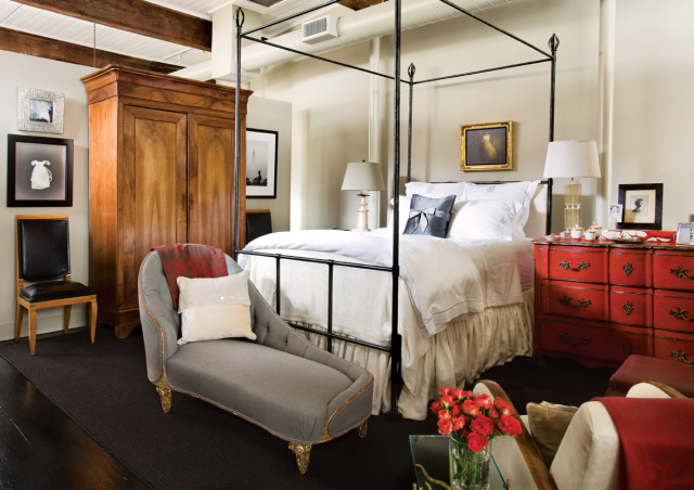 Bedroom with a four-poster bed, red ornate dresser, wooden armoire, and gray chaise lounge. White bedding and pillows with a blue accent pillow are visible.