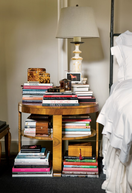 Wooden side table next to a bed, stacked with books and decorative boxes. A lamp with a cream shade sits on top.