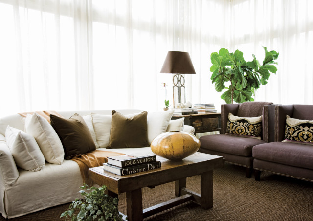 Living room with a white slipcovered sofa, brown velvet pillows, and a rustic wooden coffee table holding books and a large ceramic vase.