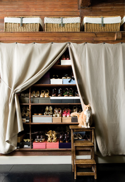 Wooden shelves filled with shoes and boxes, partially covered by a beige curtain. An orange cat sits on a wooden step stool.