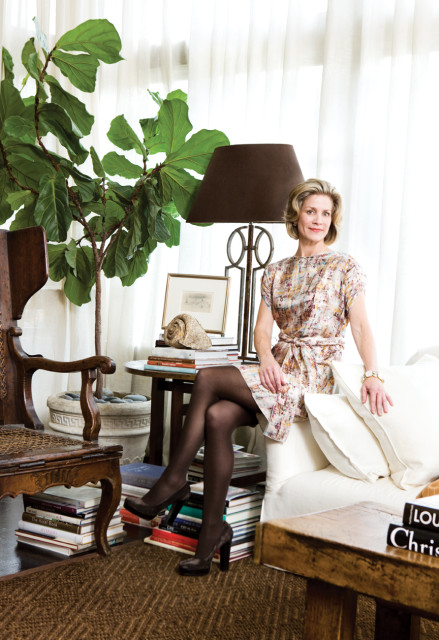 Woman in patterned dress sitting on a white sofa next to a fiddle leaf fig plant, stacked books, and a table lamp.