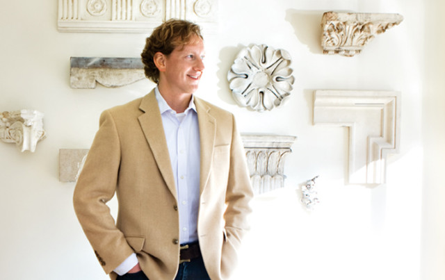 Man in a tan blazer and light blue collared shirt stands with hands in pockets in front of a white wall displaying architectural fragments.