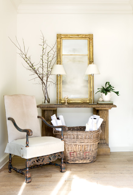 Cream upholstered armchair with carved dark wood frame next to a console table with a gold ornate mirror, two lamps, and a basket of rolled papers.