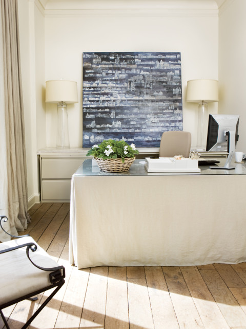 Home office with a desk covered by a cream linen skirt, two glass lamps, a computer, and a large abstract painting of buildings on the wall.