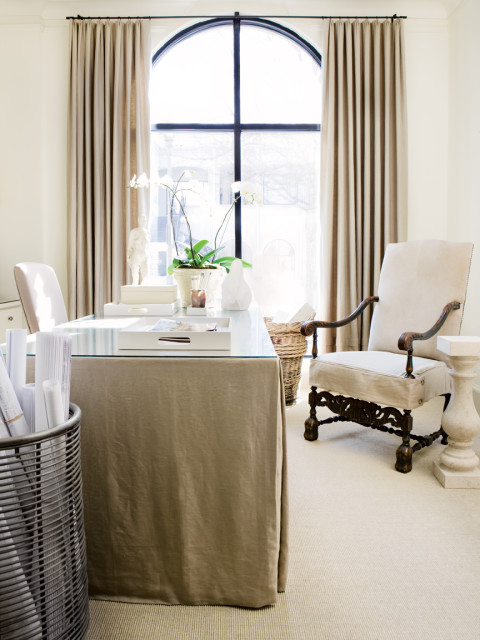 Office with a glass-top desk draped in taupe fabric, a tall arched window with taupe curtains, and a carved wooden armchair with cream upholstery.