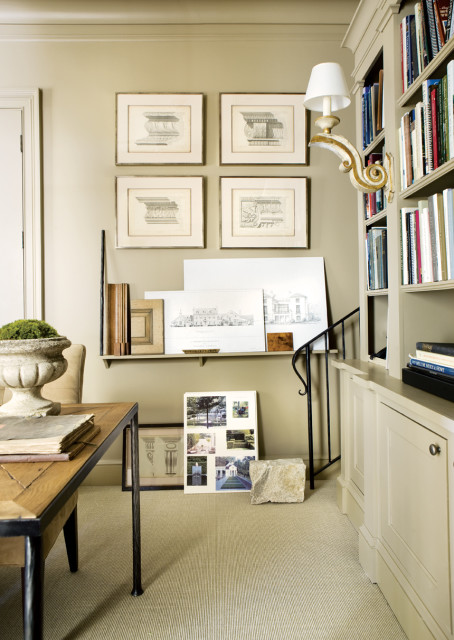 Office with tan walls, built-in bookshelves filled with books, a sconce with a white shade, and framed architectural drawings on the wall.