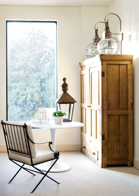 Cream carpeted room with a tall wooden cabinet, a white tulip table with a decorative birdcage, lantern, and potted plant, and a metal-framed chair with cream cushions.