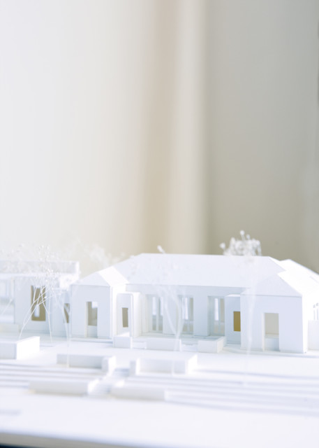 Architectural model of a white house with visible windows and doorways, surrounded by miniature trees and landscaping elements.