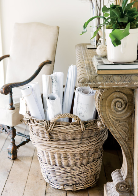 Wicker basket filled with rolled blueprints next to a carved wooden console table and upholstered armchair.