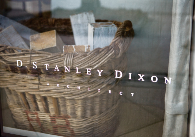 Reflection of a woven basket filled with wooden blocks on a glass surface with white text reading "D. STANLEY DIXON ARCHITECT"