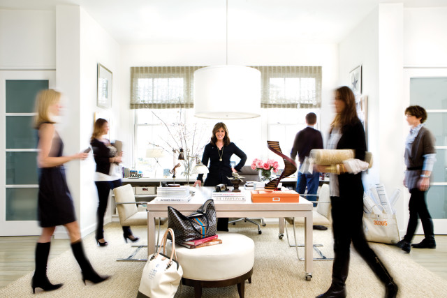 Woman in black standing behind a desk in a bright office with large windows, people blurred in motion around her.