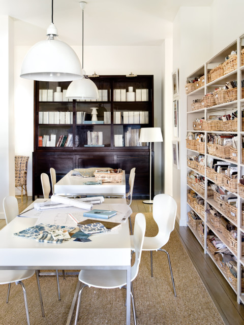 White tables with blueprints and fabric swatches in a room with a dark wood bookshelf and shelves filled with woven baskets.