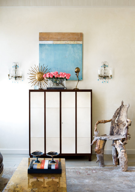 Living room with a cream wall, featuring a tall white cabinet with dark trim, a sunburst mirror, pink roses in a glass vase, and a unique driftwood chair.