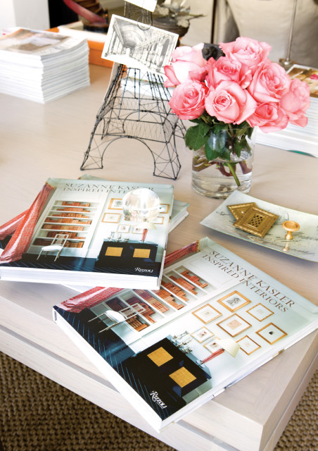 Two open books titled "Suzanne Kasler Inspired Interiors" by Rizzoli sit on a light wood table. A vase of pink roses, a wire Eiffel Tower, and decorative trays are also on the table.