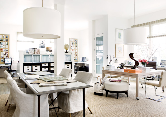 Bright office space with a long metal table and linen-covered chairs, a desk with a laptop and pink roses, and white drum pendant lights.