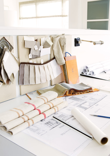 Interior design mood board with fabric swatches in neutral tones, architectural drawings, rolled blueprints, and a desk lamp.