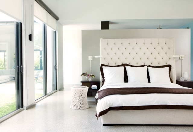 Modern bedroom with a tufted white headboard, white bedding with brown trim, and floor-to-ceiling windows overlooking a lawn.