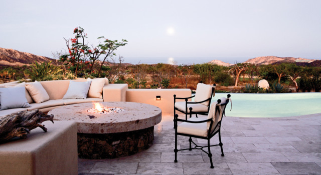 Terrace with built-in sectional sofa and stone fire pit overlooking a pool and arid landscape with mountains in the distance.