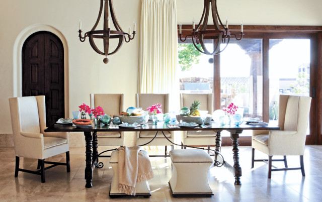 Dining room with a dark wood table set with fruit and flowers, flanked by upholstered chairs and two wooden chandeliers overhead.