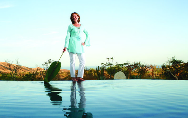 Woman in a light blue tunic and white pants stands at the edge of an infinity pool, holding a large green leaf.
