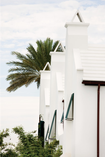 Row of white stucco buildings with triangular roof peaks and green shutters, featuring a palm tree and lush greenery against a cloudy sky.